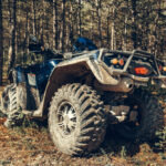 Štvorkolka so zablatenými kolesami zaparkovaná v lesnom prostredí, pripravená na jazdu v teréne.